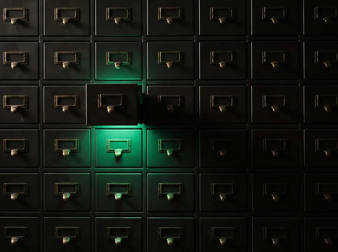 A wall of dark filing drawers, one of them open and glowing emerald